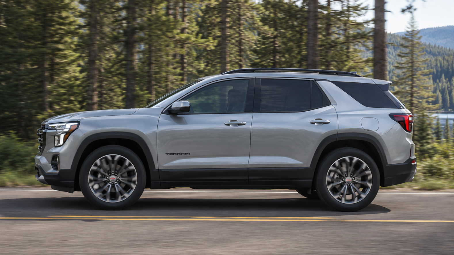 A silver GMC Terrain driving in profile by a forest.